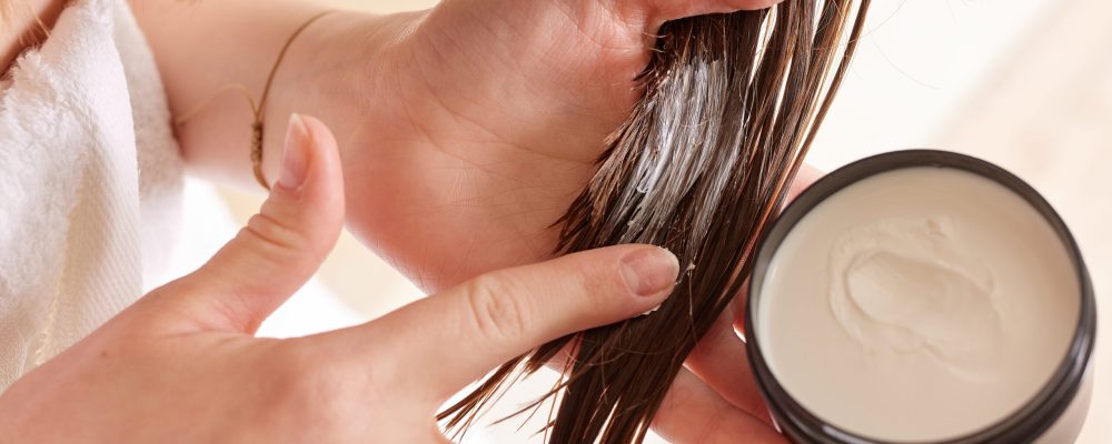 A person applying a hair treatment cream to their hair. The focus is on the hands and hair, with a jar of cream visible. The setting is bright and clean, suggesting a beauty or self-care routine.