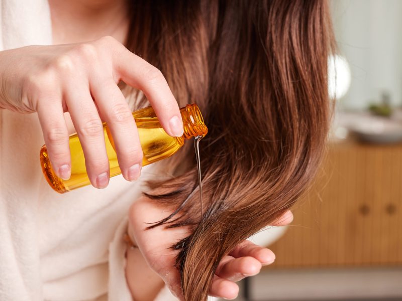 woman applying oil to her long shiny hair showing close up of hair and bottle of oil in background bright modern bathroom with soft focus background.